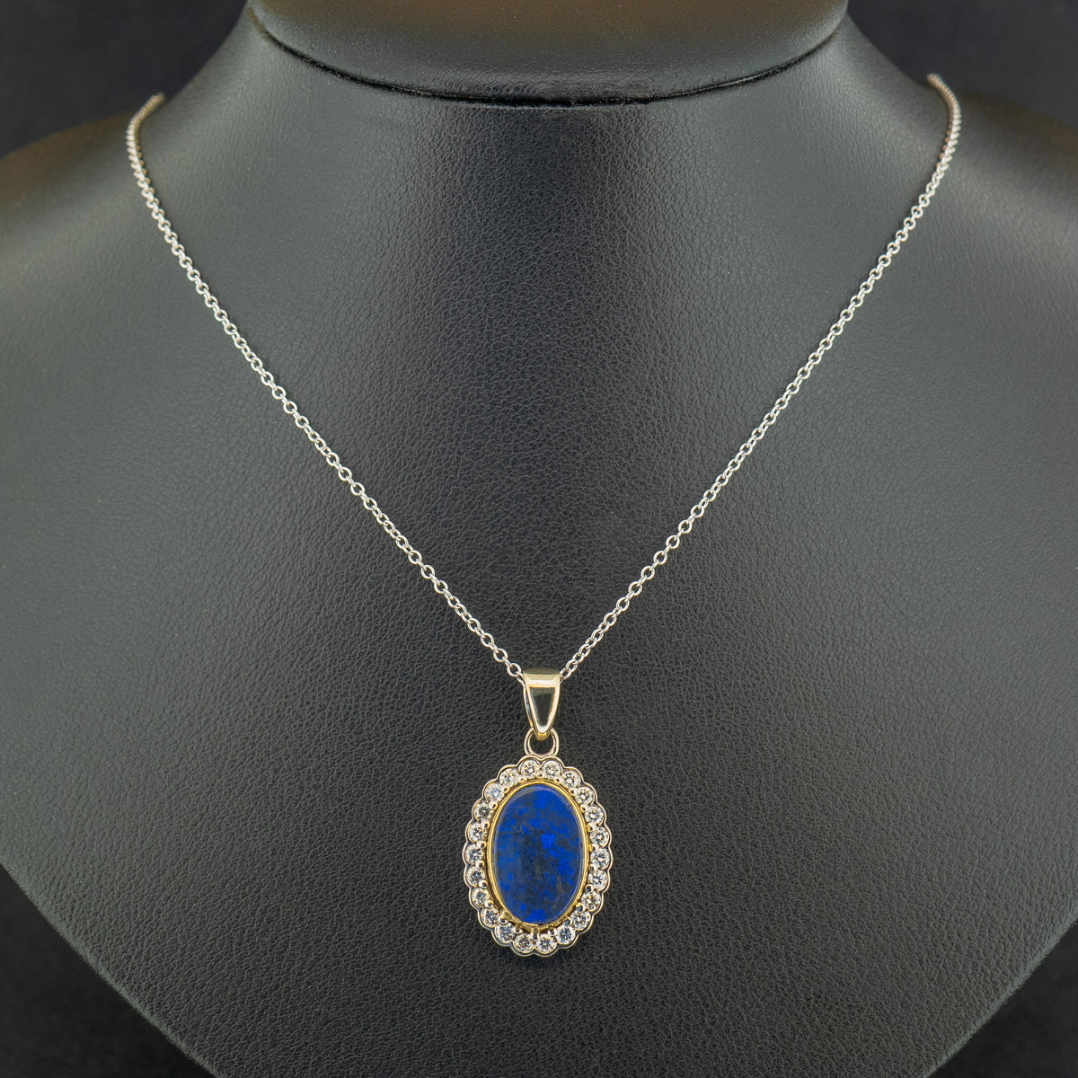 An 2.78ct Oval Blue Australian Opal with a yellow gold rubover setting and surrounded by a cluster of 0.55ct round-cut diamonds, set in white gold and on a white gold chain, hanging on the black jewellery bust.